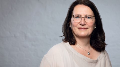 "Porträt einer Frau mittleren Alters mit schulterlangenm schwarzem Haar. Sie trägt eine Brille mit transparentem Rahmen und hat einen ruhigen, freundlichen Gesichtsausdruck. Um ihren Hals trägt sie eine silberne Kette mit einem ovalen Anhänger. In ihren Ohren sind diskrete Perlenohrringe sichtbar. Sie trägt ein beiges, cremefarbenes T-Shirt mit rundem Ausschnitt. Der Hintergrund ist neutral grau gehalten, was den Fokus auf die Person lenkt. Die Beleuchtung ist weich und gleichmäßig."