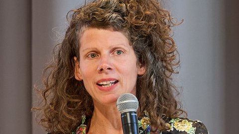 Frau mit schulterlangen, lockigen braunen Haaren, die fröhlich in ein schwarzes Mikrofon spricht. Sie trägt ein schwarzes Oberteil mit buntem Blumenmuster und einen breiten goldenen Ring. Ihr Mund ist leicht geöffnet und sie lächelt. Der Hintergrund ist unscharfgestellt in Grautönen.
