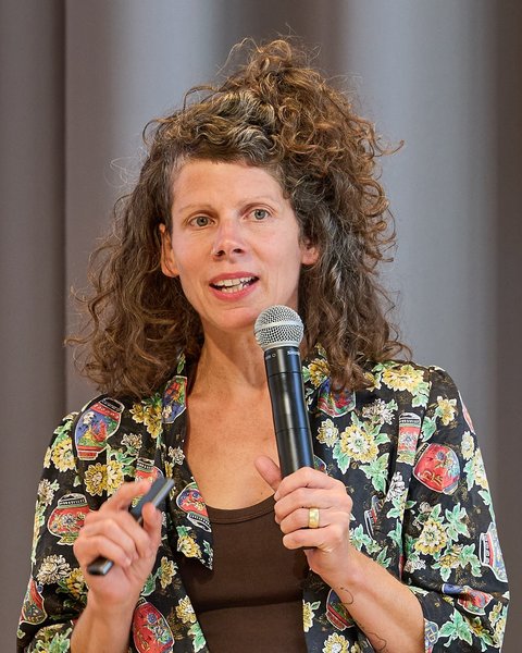 Frau mit schulterlangen, lockigen braunen Haaren, die fröhlich in ein schwarzes Mikrofon spricht. Sie trägt ein schwarzes Oberteil mit buntem Blumenmuster und einen breiten goldenen Ring. Ihr Mund ist leicht geöffnet und sie lächelt. Der Hintergrund ist unscharfgestellt in Grautönen.