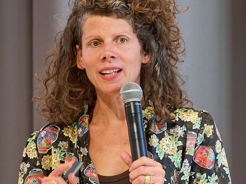 Frau mit schulterlangen, lockigen braunen Haaren, die fröhlich in ein schwarzes Mikrofon spricht. Sie trägt ein schwarzes Oberteil mit buntem Blumenmuster und einen breiten goldenen Ring. Ihr Mund ist leicht geöffnet und sie lächelt. Der Hintergrund ist unscharfgestellt in Grautönen.