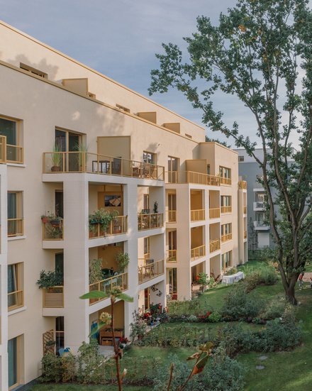 Wohnanlage am grünen Innenhof: Versetzte Loggien mit gelben Geländern, ein großer alter Baum prägt die Mitte; geschwungener Weg, Sitzbänke und gepflegte Beete, einzelne Personen nutzen den Garten.