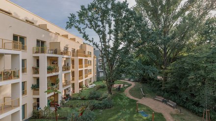 Wohnanlage am grünen Innenhof: Versetzte Loggien mit gelben Geländern, ein großer alter Baum prägt die Mitte; geschwungener Weg, Sitzbänke und gepflegte Beete, einzelne Personen nutzen den Garten.