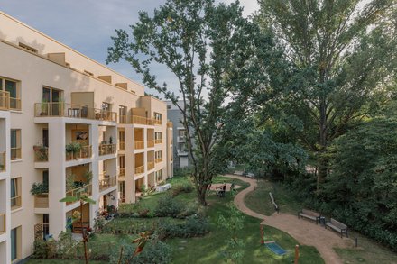 Wohnanlage am grünen Innenhof: Versetzte Loggien mit gelben Geländern, ein großer alter Baum prägt die Mitte; geschwungener Weg, Sitzbänke und gepflegte Beete, einzelne Personen nutzen den Garten.