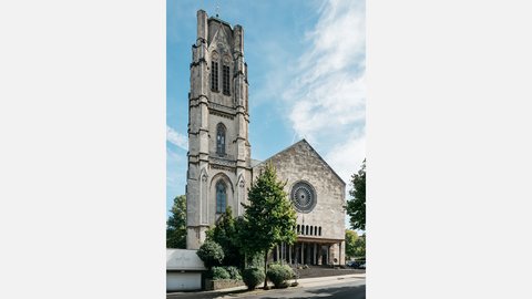 Ansicht einer Kirche mit hohem Glockenturm und heller Natursteinfassade. Vor dem Eingang stehen Bäume und Sträucher, darüber spannt sich ein blauer Himmel mit leichten Wolken.