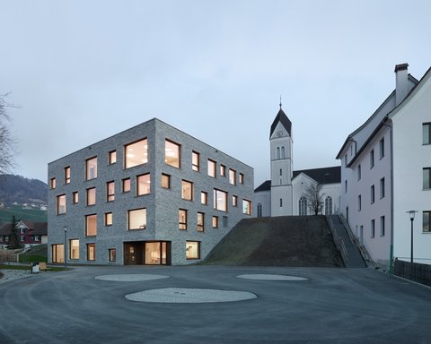 Modernes, kubisches Gebäude mit vielen quadratischen Fenstern neben einer Kirche mit Turm auf einem Hügel