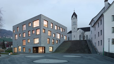 Modernes, kubisches Gebäude mit vielen quadratischen Fenstern neben einer Kirche mit Turm auf einem Hügel