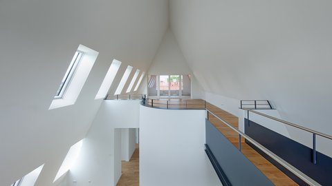 Innenraum, Galerieebene: Helles, offenes Geschoss mit hoher, spitz zulaufender Decke, Reihe schräger Dachfenster links, Holzfußboden, Metallgeländer und Brüstung, am Ende verglaste Terrassentür.