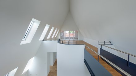 Innenraum, Galerieebene: Helles, offenes Geschoss mit hoher, spitz zulaufender Decke, Reihe schräger Dachfenster links, Holzfußboden, Metallgeländer und Brüstung, am Ende verglaste Terrassentür.