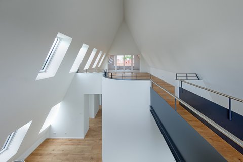 Innenraum, Galerieebene: Helles, offenes Geschoss mit hoher, spitz zulaufender Decke, Reihe schräger Dachfenster links, Holzfußboden, Metallgeländer und Brüstung, am Ende verglaste Terrassentür.