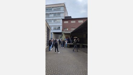 Gruppe von etwa 15–20 Menschen in einem gepflasterten Hinterhof zwischen Büro- und Lagergebäuden; eine Person vorne gestikuliert erklärend, die anderen schauen zu.