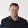 Mann mit Brille in dunkler Kleidung vor hellem Hintergrund.