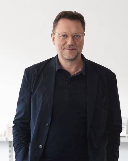 Mann mit Brille in dunkler Kleidung vor hellem Hintergrund.