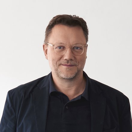 Mann mit Brille in dunkler Kleidung vor hellem Hintergrund.