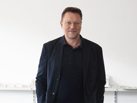 Mann mit Brille in dunkler Kleidung vor hellem Hintergrund.