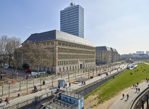 Großes historisches Gebäude mit steilem Dach vor einem hohen modernen Bürogebäude und einem Park mit Spaziergängern und Sitzenden