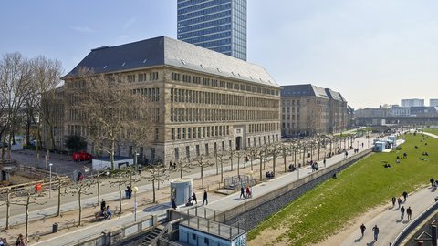 Großes historisches Gebäude mit steilem Dach vor einem hohen modernen Bürogebäude und einem Park mit Spaziergängern und Sitzenden