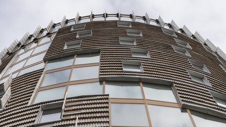 Fassadendetail von unten: Blick in die vertikale Fassade des Turms mit reliefartiger Bekleidung aus kleinformatigen, strukturierten Platten, schmalen Fensteröffnungen und bronzefarbenen Fensterprofilen, perspektivisch nach oben ziehend.