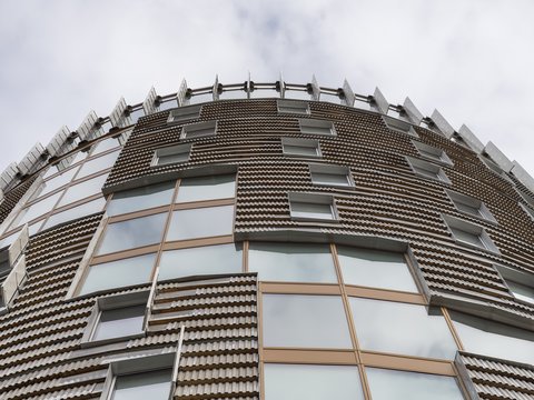 Fassadendetail von unten: Blick in die vertikale Fassade des Turms mit reliefartiger Bekleidung aus kleinformatigen, strukturierten Platten, schmalen Fensteröffnungen und bronzefarbenen Fensterprofilen, perspektivisch nach oben ziehend.