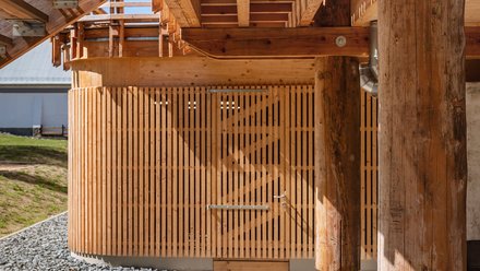 Konstruktionsdetail unter dem Steg: schwere Holzträger, Rundholzstützen auf Metallfüßen, gebogene Lamellenwand mit Tor.