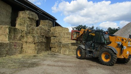 Hof: Gelber Teleskoplader stapelt rechteckige Strohballen vor einer Scheune.