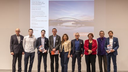 Eine Gruppe von Erwachsenen steht nebeneinander auf einer Bühne vor einer Projektion der Brücke, einige halten Broschüren in den Händen und blicken in die Kamera, wodurch das Bild eine ausgezeichnete Projektgemeinschaft dokumentiert und für Teamarbeit, interdisziplinäre Zusammenarbeit und öffentliche Würdigung von Baukultur steht.