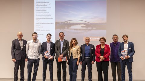 Eine Gruppe von Erwachsenen steht nebeneinander auf einer Bühne vor einer Projektion der Brücke, einige halten Broschüren in den Händen und blicken in die Kamera, wodurch das Bild eine ausgezeichnete Projektgemeinschaft dokumentiert und für Teamarbeit, interdisziplinäre Zusammenarbeit und öffentliche Würdigung von Baukultur steht.