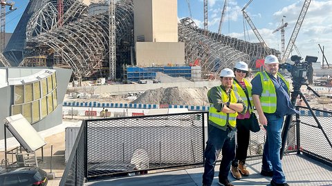 "Vier Personen stehen auf einer erhöhten Baustellen-Plattform mit schwarzem Metallgeländer. Drei tragen weiße Schutzhelme und gelb-grüne Warnwesten. Von links nach rechts: ein Mann in grüner Weste, eine Frau in rosa und gelber Weste in der Mitte, und rechts zwei Männer in gelber und blauer Weste. Die Baustelle zeigt massive Stahlkonstruktionen, mehrere rote und gelbe Baukräne, beige Betonblöcke und hohe Metallgerüste unter blauem Himmel. Links im Hintergrund ein moderner Gebäudeteil mit gelben Paneelen. Eine stationäre Filmkamera auf Stativ ist rechts zu sehen."