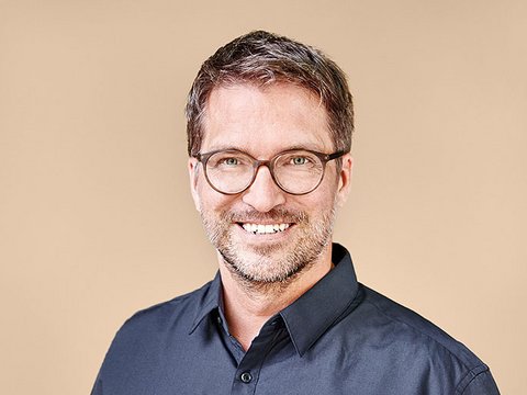 „Porträt eines lächelnden Mannes mit kurzen braunen Haaren, Bart und runder Brille. Er trägt ein dunkelblaues Hemd und steht vor einem beigefarbenen Hintergrund.“