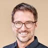 „Porträt eines lächelnden Mannes mit kurzen braunen Haaren, Bart und runder Brille. Er trägt ein dunkelblaues Hemd und steht vor einem beigefarbenen Hintergrund.“
