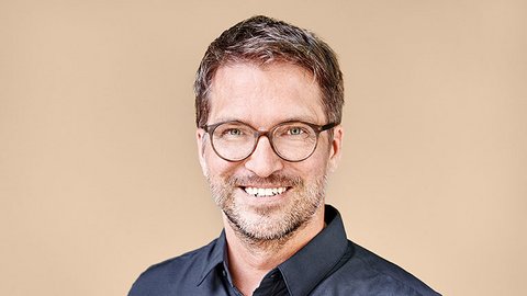„Porträt eines lächelnden Mannes mit kurzen braunen Haaren, Bart und runder Brille. Er trägt ein dunkelblaues Hemd und steht vor einem beigefarbenen Hintergrund.“