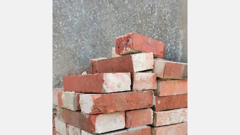 Stapel gebrauchter roter Backsteine mit Mörtelresten vor rauer Betonwand; einige Steine sind an den Kanten beschädigt.