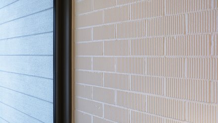 Detail: Weiß gestrichene Ziegelwand mit feinen vertikalen Rillen; links eine Fensterlaibung mit dunklem Rahmen und außenliegender, horizontal gerillter Fassade.