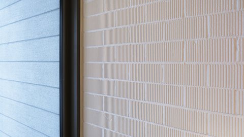 Detail: Weiß gestrichene Ziegelwand mit feinen vertikalen Rillen; links eine Fensterlaibung mit dunklem Rahmen und außenliegender, horizontal gerillter Fassade.