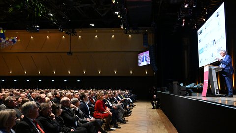 Großer Konferenzsaal: rechts spricht eine Person am Rednerpult; links sitzt ein voll besetztes Publikum. Auf der Leinwand Sponsoren‑Logos; Event „Jahresempfang der Wirtschaft“.