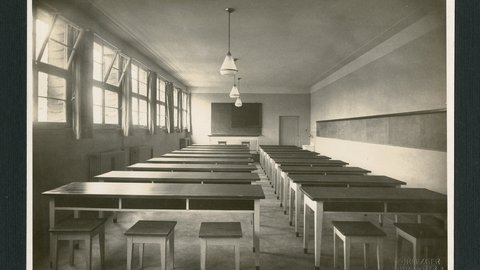 Leerer Klassenraum mit Reihen von Tischen und Hockern, großen Fenstern links und einer Tafel vorne