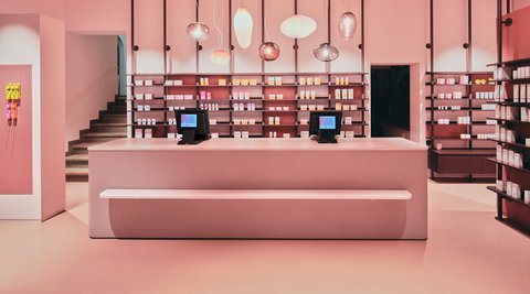 „Ein modernes Geschäft, das komplett in Pastellpink gehalten ist. Im Zentrum steht ein langer Tresen mit zwei Kassensystemen, dahinter befinden sich deckenhohe, dunkelrote Regale mit zahlreichen Produkten.“