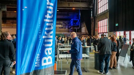 Infostand „Baukultur“ in Veranstaltungshalle Besucher:innen informieren sich an Ständen in einer großen Industriehalle.
