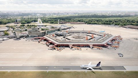 Eine Luftaufnahme zeigt den Flughafen Berlin-Tegel mit seinem markanten, hexagonalen Terminalgebäude im Zentrum. Das rote Dach des Gebäudes umschließt einen Innenhof und ist von zahlreichen Parkpositionen für Flugzeuge und Passagierbrücken umgeben. Rechts unten auf der Startbahn steht ein Flugzeug der Lufthansa. Im Hintergrund erstreckt sich eine weite, grüne Parklandschaft und dahinter die Skyline von Berlin unter einem leicht bewölkten Himmel. Der Kontrollturm ist links neben dem Terminal zu erkennen.