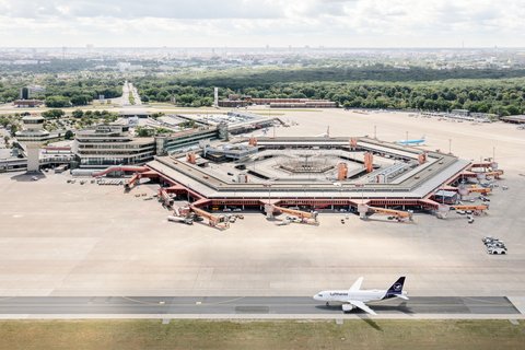 Eine Luftaufnahme zeigt den Flughafen Berlin-Tegel mit seinem markanten, hexagonalen Terminalgebäude im Zentrum. Das rote Dach des Gebäudes umschließt einen Innenhof und ist von zahlreichen Parkpositionen für Flugzeuge und Passagierbrücken umgeben. Rechts unten auf der Startbahn steht ein Flugzeug der Lufthansa. Im Hintergrund erstreckt sich eine weite, grüne Parklandschaft und dahinter die Skyline von Berlin unter einem leicht bewölkten Himmel. Der Kontrollturm ist links neben dem Terminal zu erkennen.