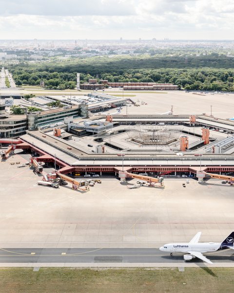 Eine Luftaufnahme zeigt den Flughafen Berlin-Tegel mit seinem markanten, hexagonalen Terminalgebäude im Zentrum. Das rote Dach des Gebäudes umschließt einen Innenhof und ist von zahlreichen Parkpositionen für Flugzeuge und Passagierbrücken umgeben. Rechts unten auf der Startbahn steht ein Flugzeug der Lufthansa. Im Hintergrund erstreckt sich eine weite, grüne Parklandschaft und dahinter die Skyline von Berlin unter einem leicht bewölkten Himmel. Der Kontrollturm ist links neben dem Terminal zu erkennen.