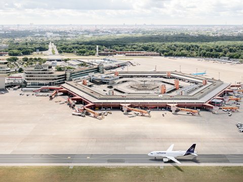 Eine Luftaufnahme zeigt den Flughafen Berlin-Tegel mit seinem markanten, hexagonalen Terminalgebäude im Zentrum. Das rote Dach des Gebäudes umschließt einen Innenhof und ist von zahlreichen Parkpositionen für Flugzeuge und Passagierbrücken umgeben. Rechts unten auf der Startbahn steht ein Flugzeug der Lufthansa. Im Hintergrund erstreckt sich eine weite, grüne Parklandschaft und dahinter die Skyline von Berlin unter einem leicht bewölkten Himmel. Der Kontrollturm ist links neben dem Terminal zu erkennen.