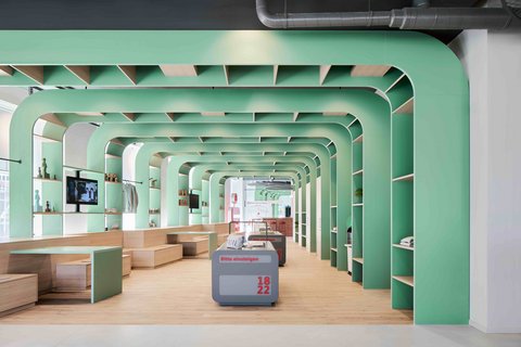 Moderner Ausstellungsraum mit mintgrünen Rundbögen, Holzpodesten und offenen Regalen.
