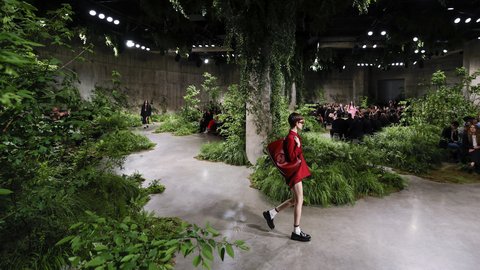 "Modenschau in einer großen Halle mit betonnen Wänden, die mit üppiger Vegetation gestaltet ist. Ein geschwungener Laufsteg führt durch Pflanzenbeete und kleine Bäume. Ein Model in rotem Outfit mit passender Tasche läuft im Vordergrund über den Weg. Weitere Models und Zuschauer sitzen entlang des Laufstegs. Hängende Pflanzen und Deckenstrahler erzeugen eine stimmungsvolle, natürliche Atmosphäre."
