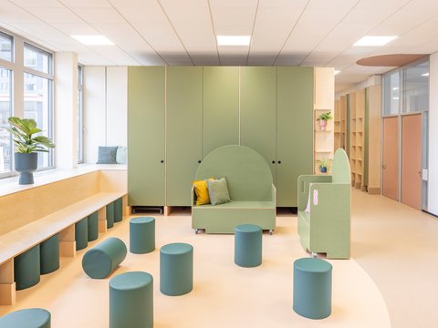 "Offener Kinderbereich mit modernem Design. Links an der Fensterfront eine Holzbank mit grünen Untersätzen. Zentral grüne Sitznische mit gelbem Kissen und grünen Kissen. Mehrere türkis-grüne zylindrische Sitzelemente verteilt im Raum. Rechts grüne Wandelemente mit abgerundeten Formen. Helle Holzböden, große Fenster mit Blick nach außen, weiße Decke mit Beleuchtung."