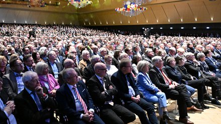 Überblick über den voll besetzten Saal; hunderte Gäste blicken zur Bühne. Bunte Ballon‑Dekorationen hängen von der Decke.