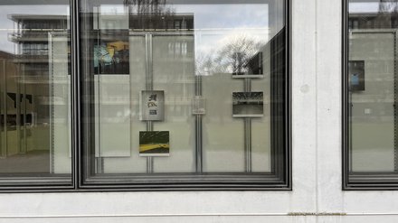 Man sieht eine Glasfassade von außen. Hinter dem Fenster hängen mehrere Fotografien und Texttafeln an metallischen Trägern. Spiegelungen von Bäumen und Gebäuden überlagern die Ausstellungsinhalte. Das Bild vermittelt, dass die Ausstellung auch nach außen sichtbar ist. Innen- und Außenraum, Realität und Reflexion überlagern sich – ähnlich wie physische Welt und digitale Sphäre.