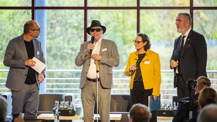 Vier Personen stehen vor einer Fensterfront auf einer Bühne und sprechen zu einem Publikum 