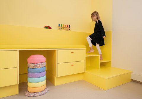 „Ein Mädchen klettert auf die Stufen eines leuchtend gelben Einbaumöbels mit integriertem Schreibtisch. Im Vordergrund steht ein Turm aus bunten, ringförmigen Kissen.“