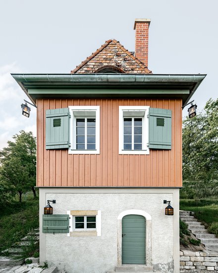 Ein kleines, freistehendes Haus mit rechteckigem Grundriss steht am Hang in einer grünen Umgebung, der untere Teil ist verputzt und massiv, der obere Teil besteht aus vertikal verschalten Holzflächen in warmem Farbton, grüne Fensterläden, ein steiles Ziegeldach und ein gemauerter Schornstein prägen das Erscheinungsbild, eine Außentreppe führt seitlich nach oben, wodurch das Bild ein sorgfältig instand gesetztes oder neu interpretiertes Weinberg- oder Gartenhaus zeigt, das traditionelle Bauweise mit zeitgemäßer Nutzung verbindet und für den behutsamen Umgang mit kleinteiliger historischer Architektur und Landschaft steht.