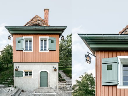 Ein kleines, freistehendes Haus mit rechteckigem Grundriss steht am Hang in einer grünen Umgebung, der untere Teil ist verputzt und massiv, der obere Teil besteht aus vertikal verschalten Holzflächen in warmem Farbton, grüne Fensterläden, ein steiles Ziegeldach und ein gemauerter Schornstein prägen das Erscheinungsbild, eine Außentreppe führt seitlich nach oben, wodurch das Bild ein sorgfältig instand gesetztes oder neu interpretiertes Weinberg- oder Gartenhaus zeigt, das traditionelle Bauweise mit zeitgemäßer Nutzung verbindet und für den behutsamen Umgang mit kleinteiliger historischer Architektur und Landschaft steht.